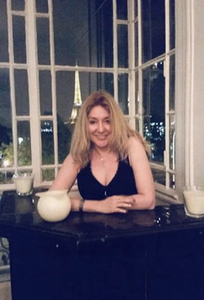 Photo de Stady du Lac, une médium à Paris, blonde et souriante, accoudé à une table devant une grande fenêtre ancienne par laquelle on aperçoit la Tour Eiffel